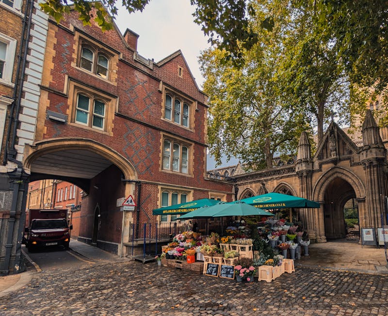 An outdoor flower stall situated on a cobblestone street beneath a large green umbrella, featuring a vibrant display of fresh flowers including pink, yellow, red, and white blooms arranged in bouquets