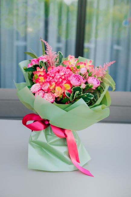A vibrant floral bouquet featuring predominantly pink flowers, including roses, pink gypsophila, and other mixed blooms, arranged in a lush, rounded style. The bouquet is wrapped in light green tissue