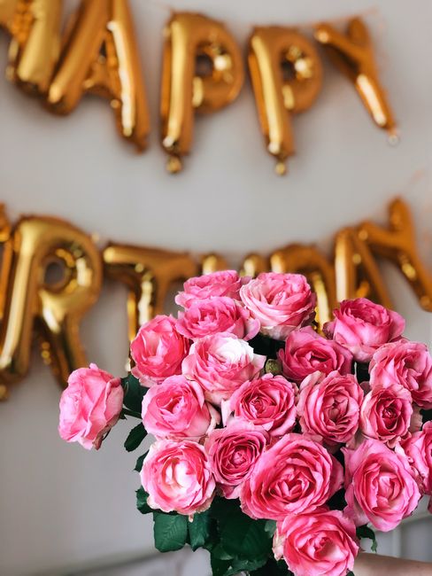 A vibrant bouquet of fresh pink roses with lush green leaves, displayed in front of gold foil balloons spelling out 'HAPPY BIRTHDAY'. The roses are tightly packed, showcasing their layered petals with