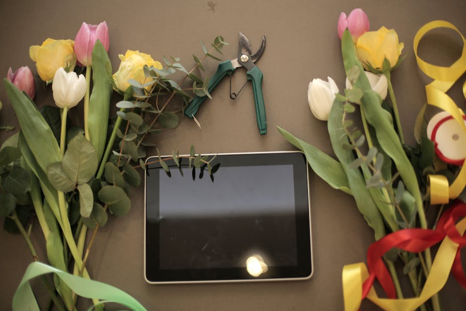 A flat lay of fresh flower bouquets featuring pink, yellow, and white tulips with green leaves and eucalyptus stems, arranged on a neutral-colored surface. In the center, there is a black tablet with