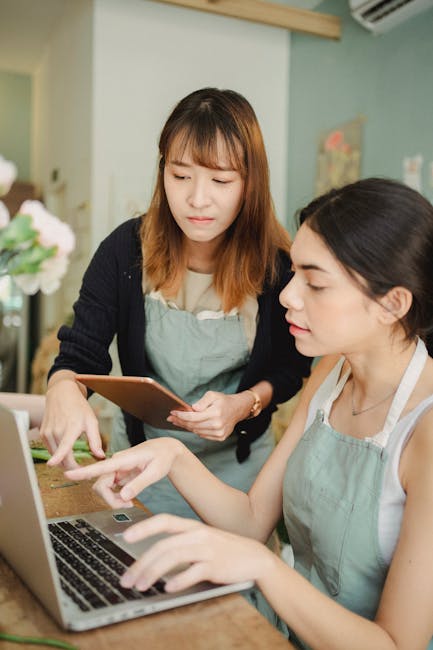 The image shows two women in a florist shop or flower arranging studio. One woman, with shoulder-length light brown hair and wearing a beige top and light green apron, is sitting at a wooden table, us