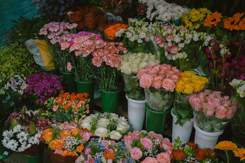 A vibrant display of fresh flowers arranged in various vases and buckets, including pink and white roses, orange and yellow blooms, purple daisies, white daisies, and other colorful floral varieties.