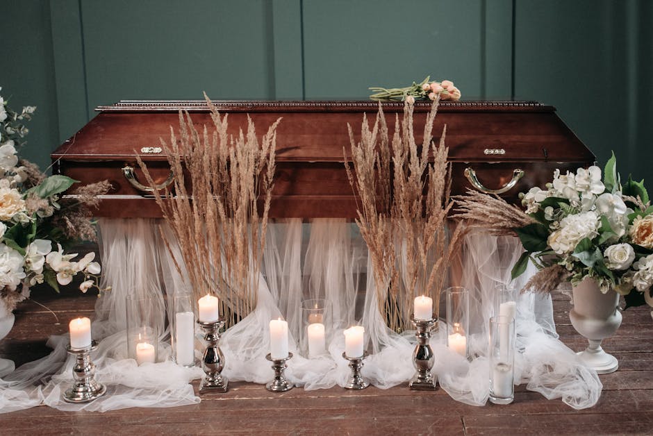 A wooden casket is decorated with a floral arrangement consisting of white roses, orchids, and pale pink flowers, accented by lush green leaves. Surrounding the casket are tall, beige dried grass stem