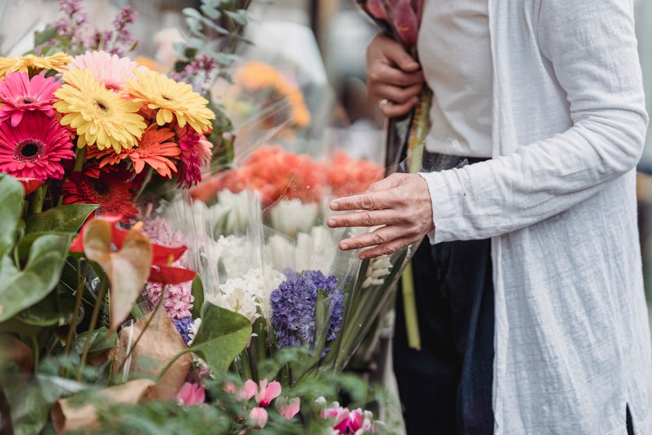 A person dressed in a white cardigan and dark pants is selecting a floral arrangement at a busy flower market, with an array of fresh, vibrant spring flowers including pink, yellow, orange, purple, an